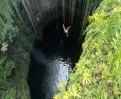 This man geared up to dive off from the top of a beautiful, green cliff into the clear pool of water below. After bracing himself, he took the plunge towards the water while people around him cheered him on.